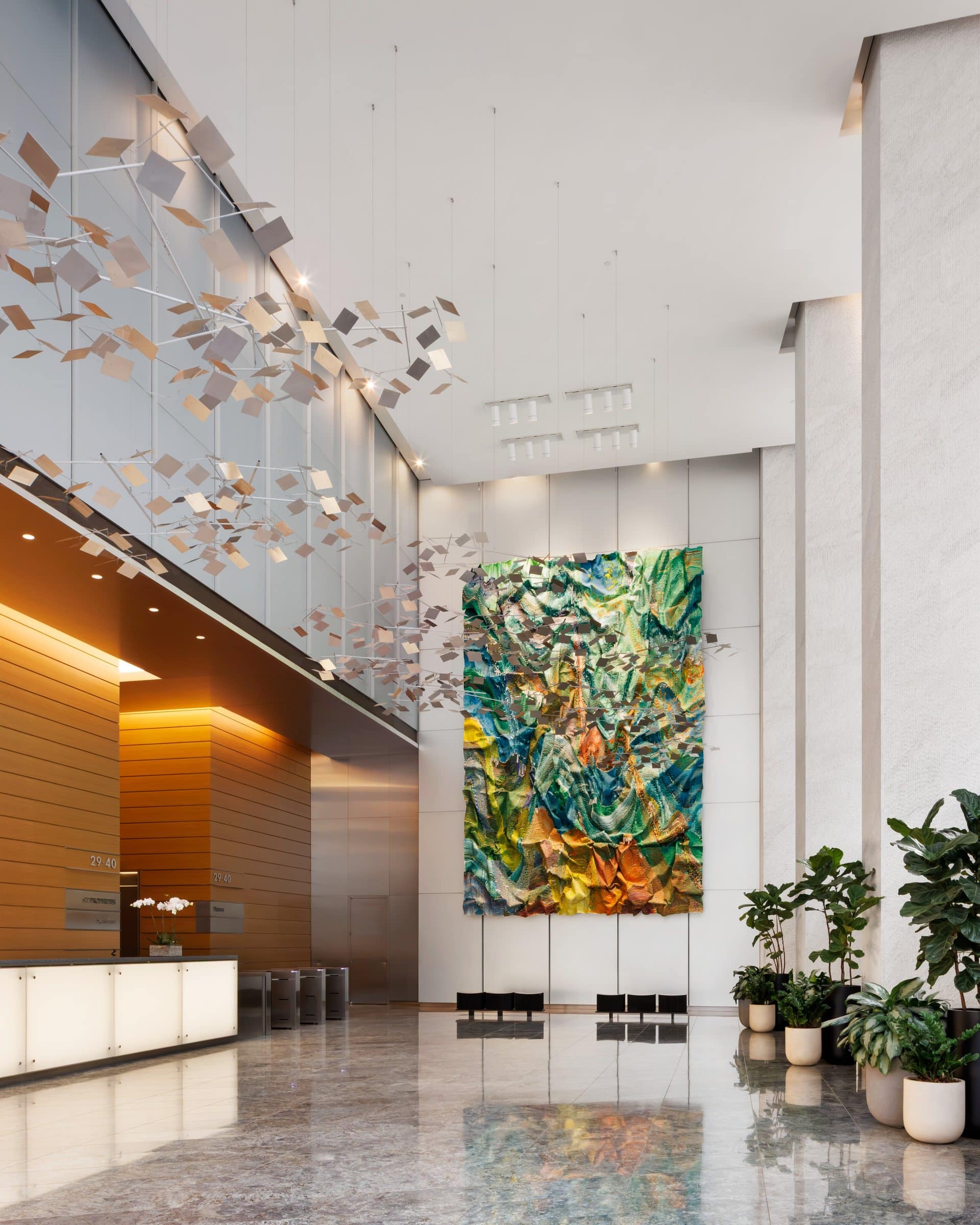 Angled lobby view toward a tall green and gold textile work, with potted plants and a reflective stone floor