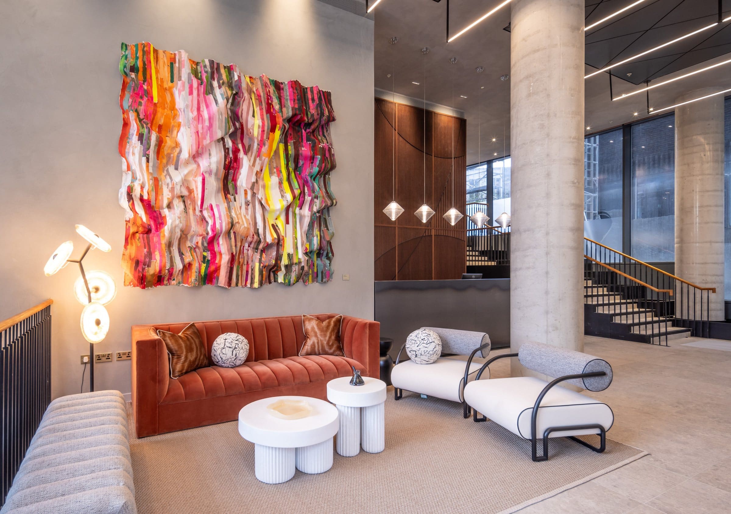 Multicolored hanging textile work above a rust sofa and pale chairs in a concrete-columned lobby