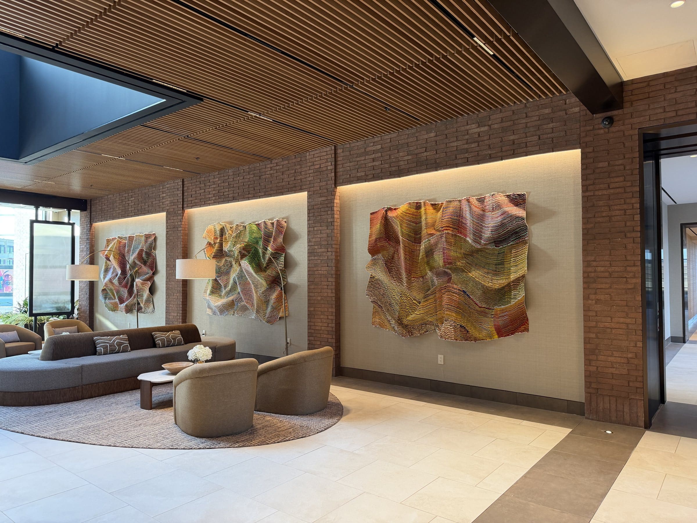 Three rippling multicolored textile works in illuminated brick-wall recesses beside lobby seating