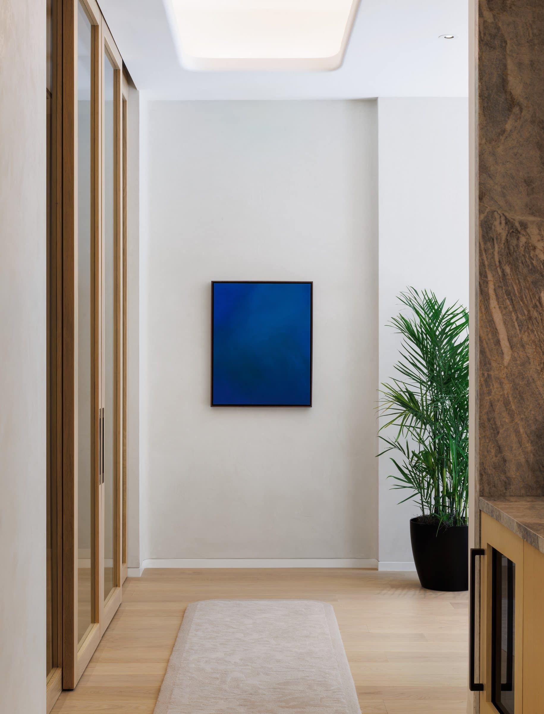 A deep blue color-field painting at the end of a pale corridor beside a potted palm