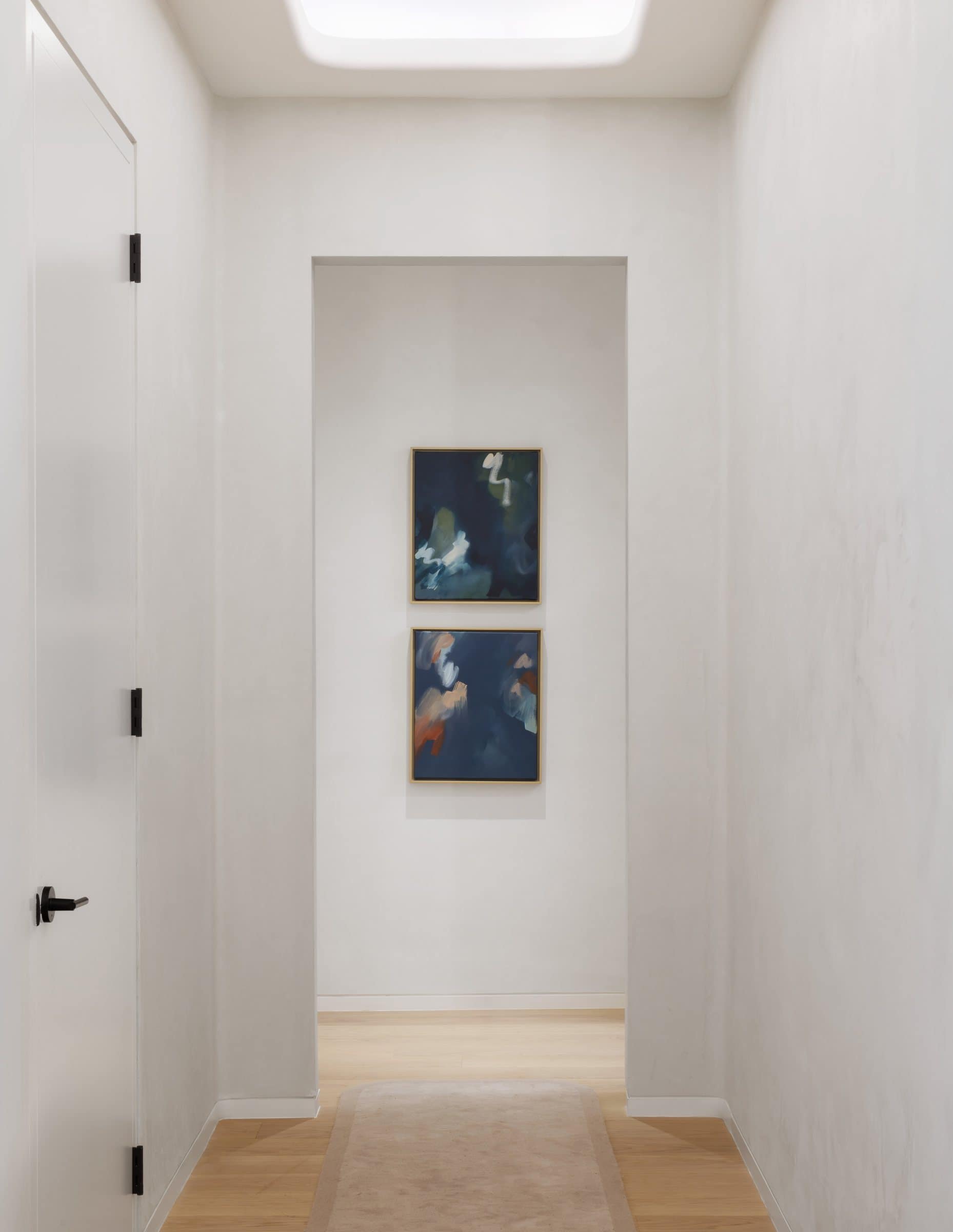 Two small dark blue-green paintings with pale brushstrokes hang vertically at the end of a white corridor