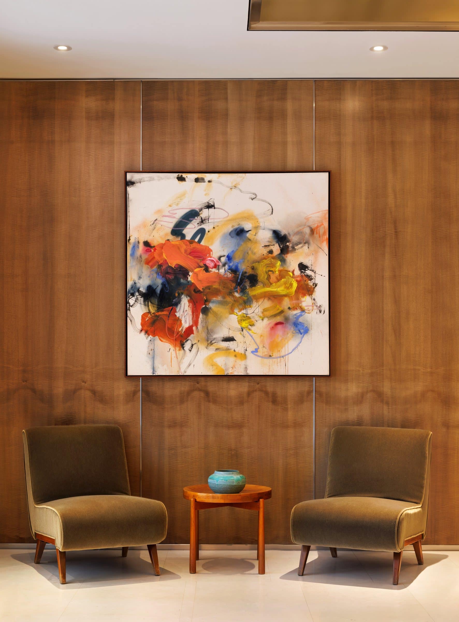 Square orange, yellow, blue and black abstract painting on an oak-paneled wall above two velvet chairs