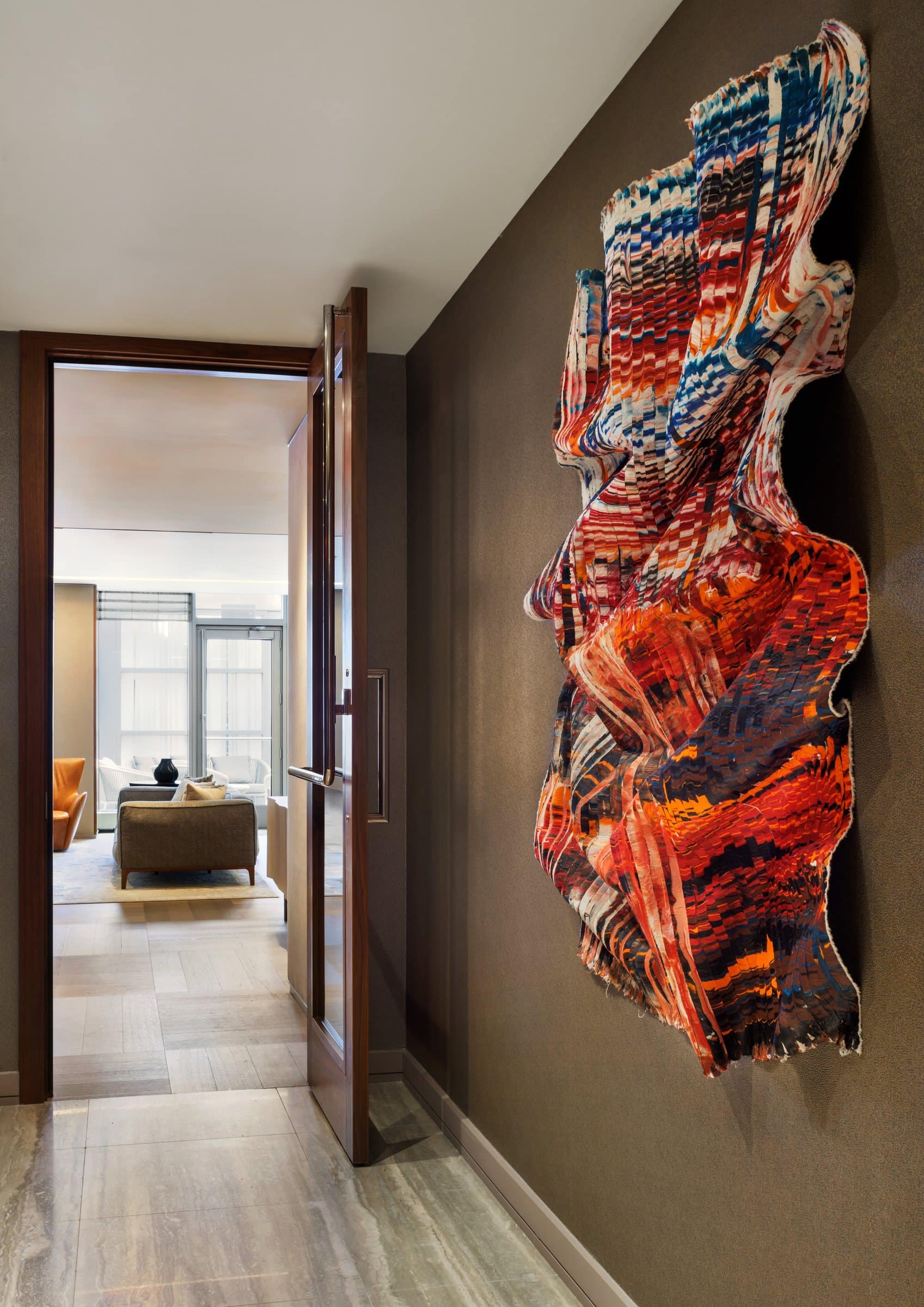 Angled close view of a red, orange, blue and white cut-silk wall work in a corridor beside an open lounge doorway