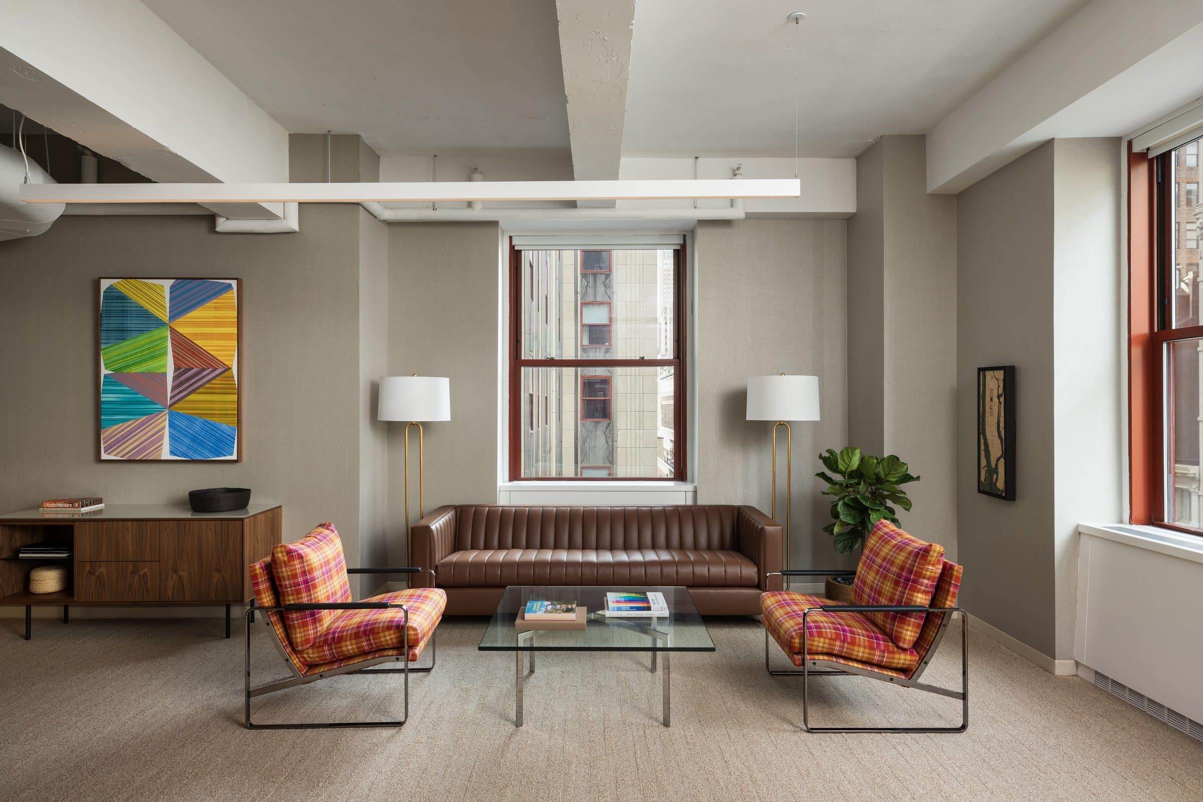 Geometric painting of striped triangles in blue, green and yellow above a walnut credenza in a lounge with plaid armchairs