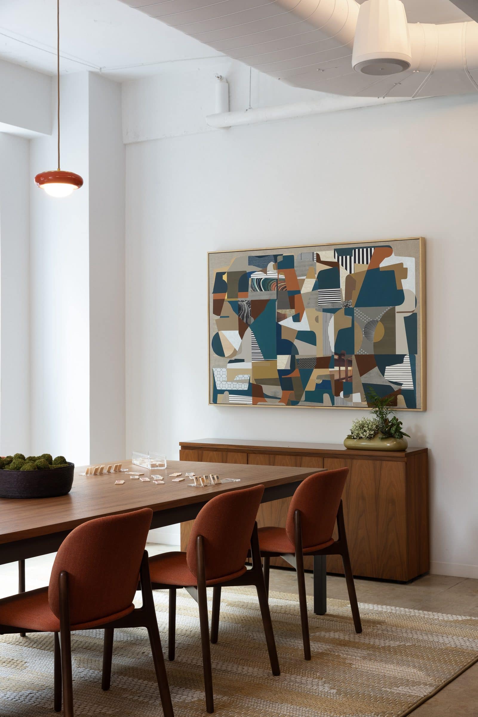 Collaged abstract painting of patterned shapes in teal, rust and cream above a credenza at the end of a long table