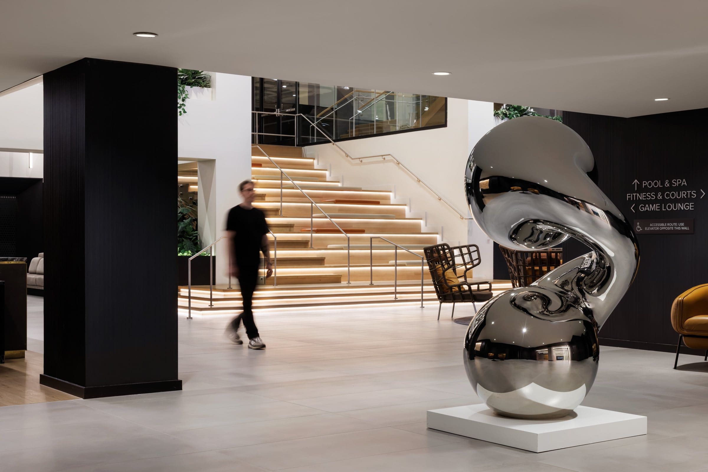 Reflective stacked sculptural forms on a white plinth beside the illuminated amenity stair, with a passing visitor