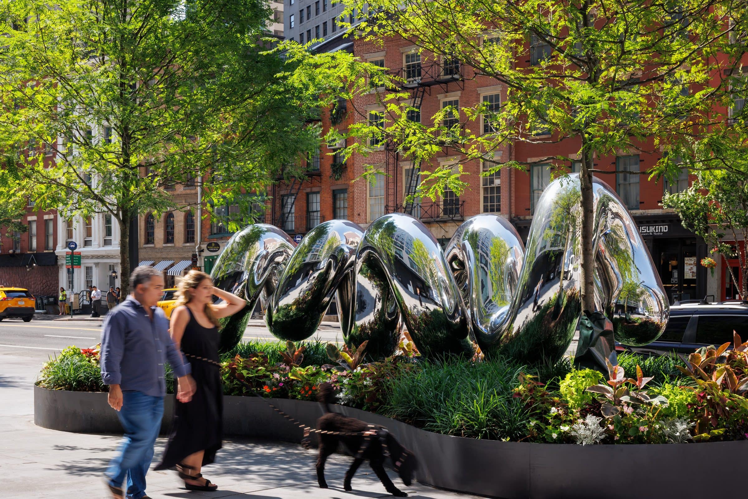 Pedestrians and a dog pass the reflective looping Love sculpture among trees and planting
