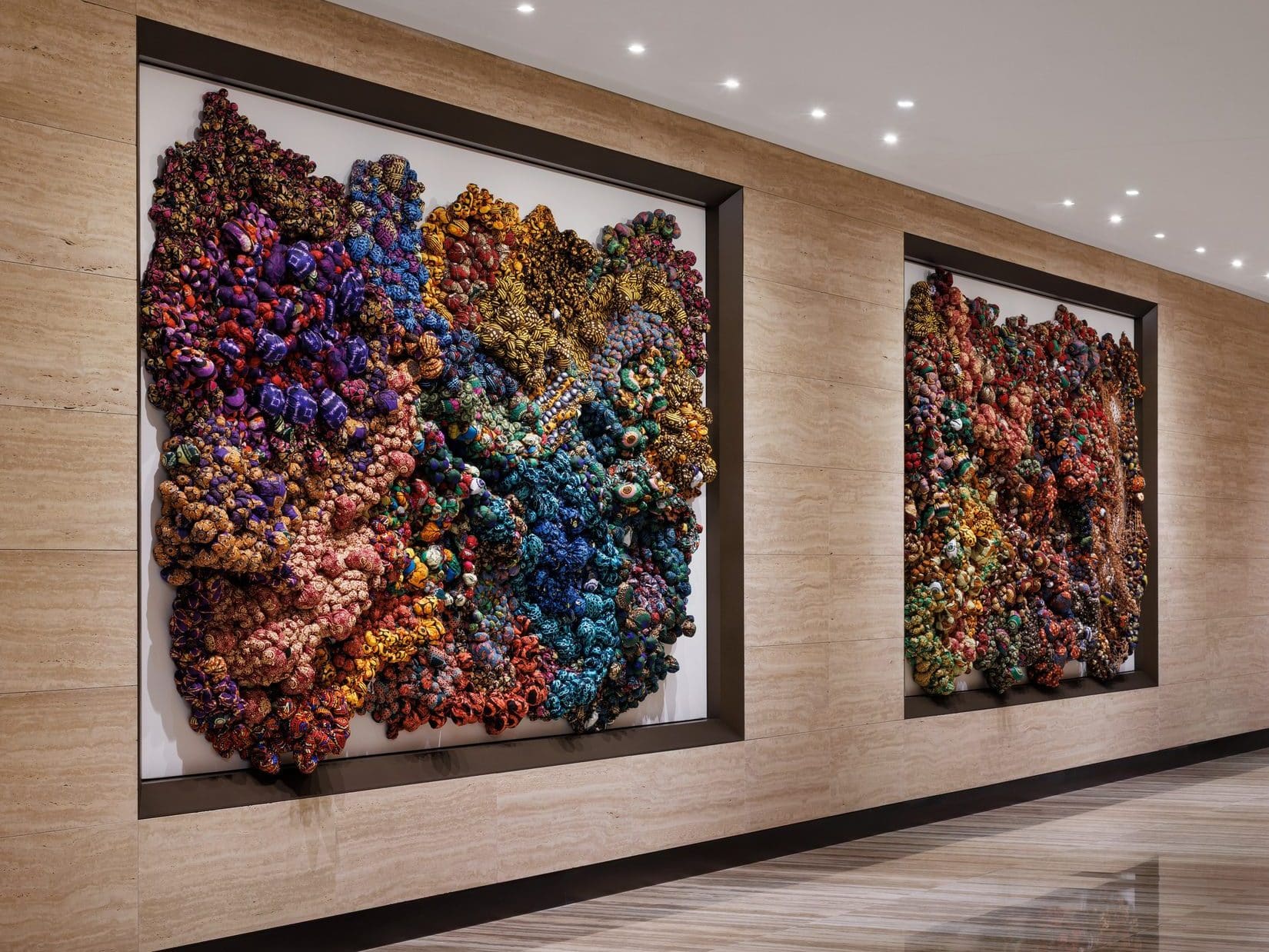 Two framed textile works of densely knotted fabric in purple, blue, gold and rust, set into a travertine corridor wall