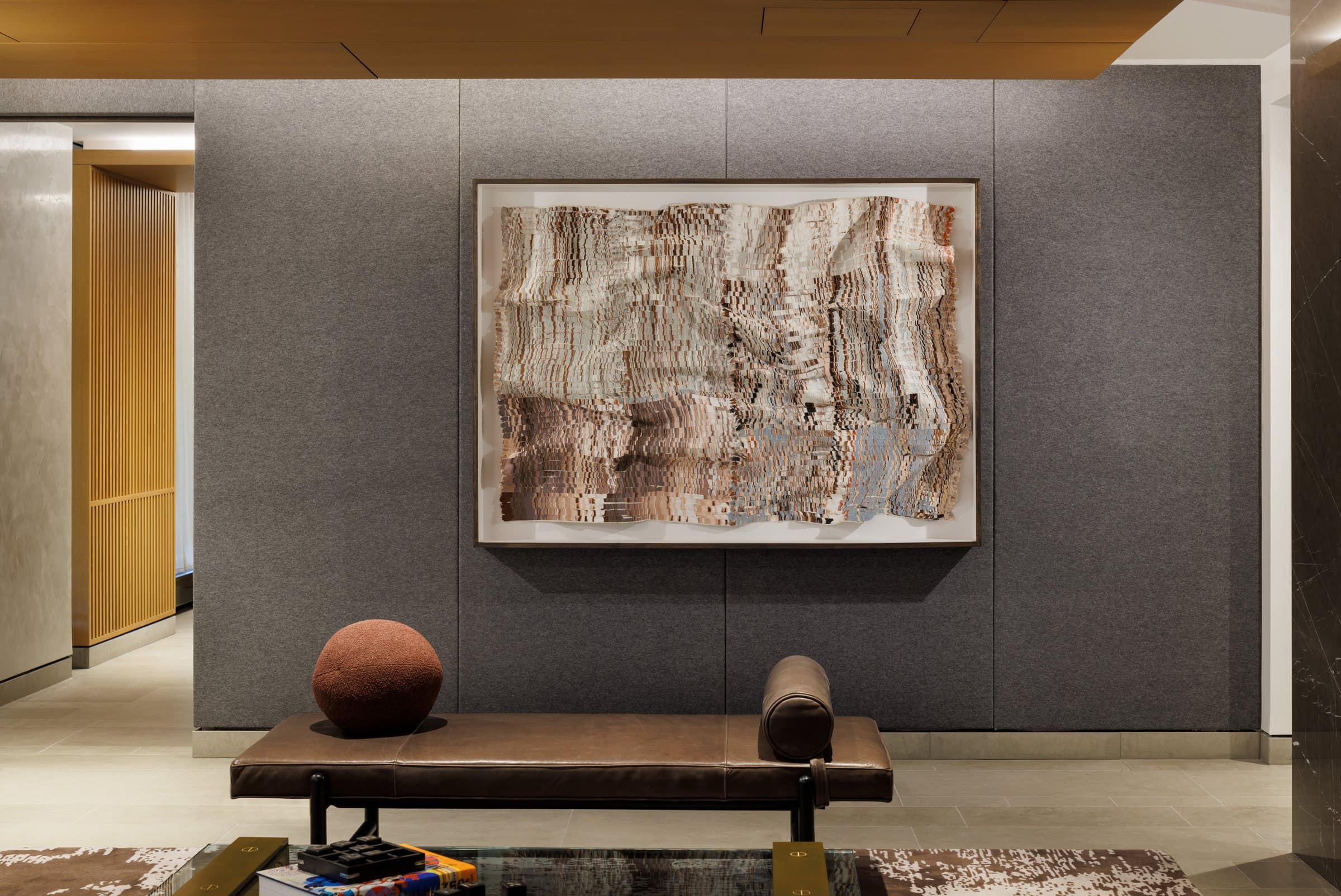 Framed rippling textile work in cream, brown and pale blue above a bench against gray wall panels