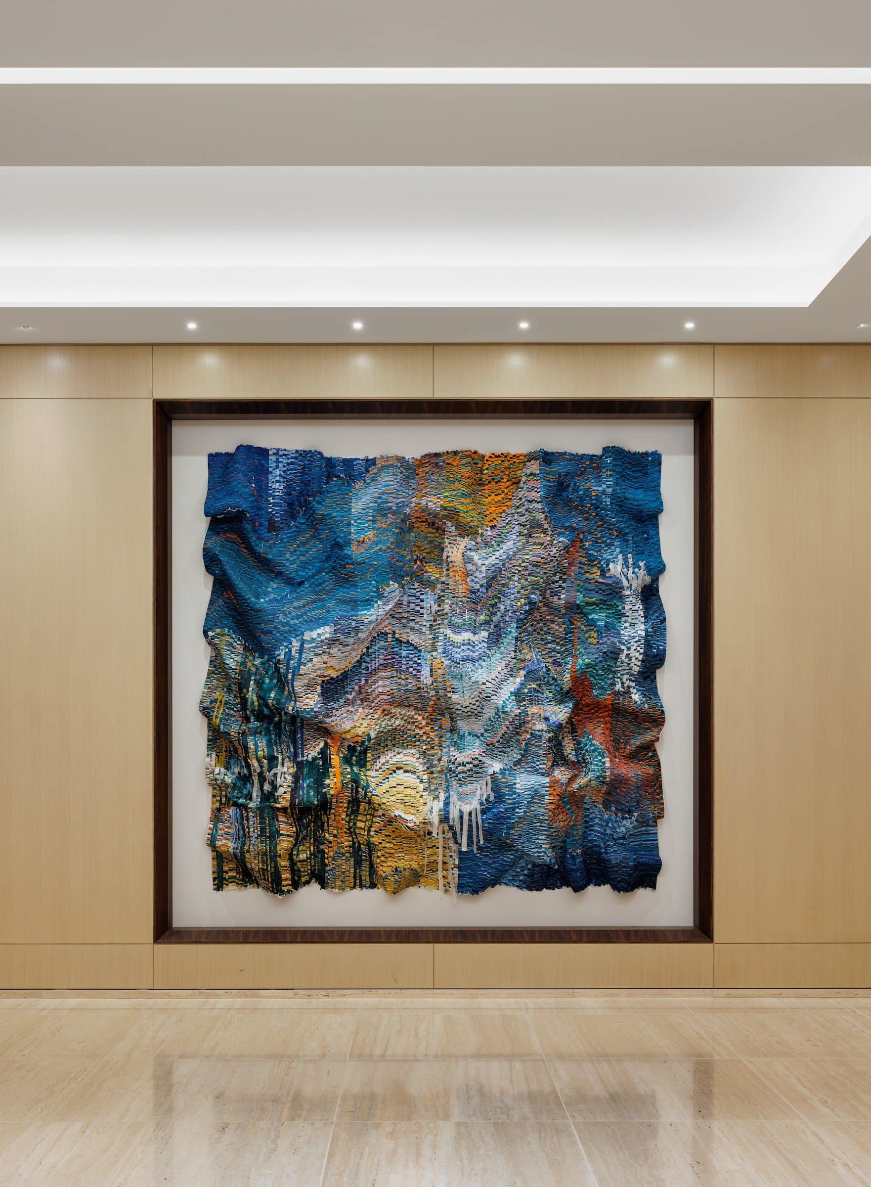 Blue, gold and orange cut-silk wall work in a dark frame within a pale wood-paneled sky-lobby niche