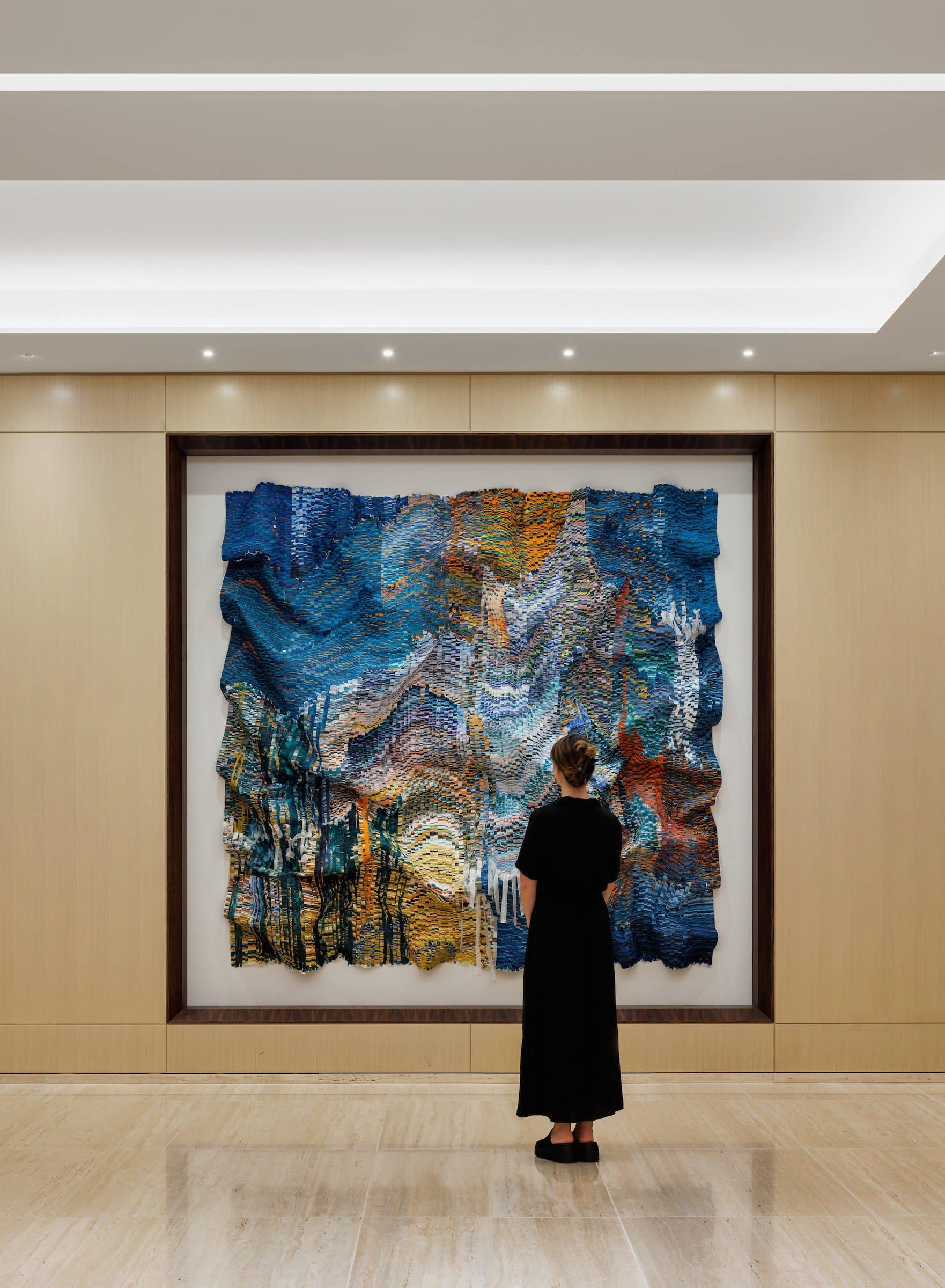 A visitor stands facing the blue-and-gold textile work in a pale wood-paneled niche