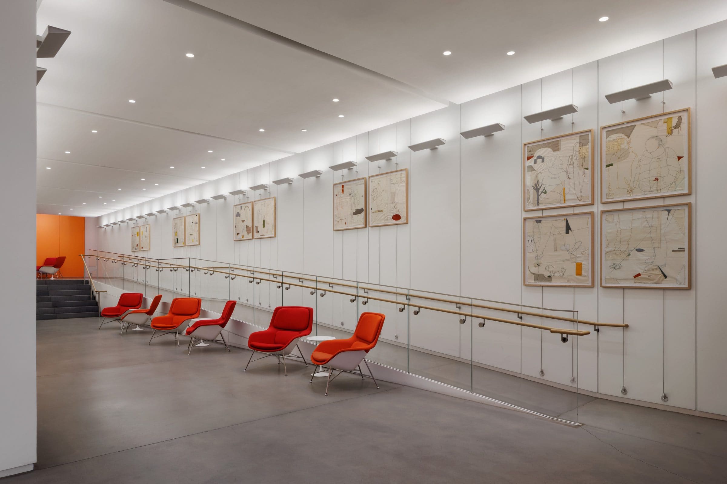 Twelve framed works of stitched line drawings and fabric on canvas line a lobby wall above a ramp, with red lounge chairs below