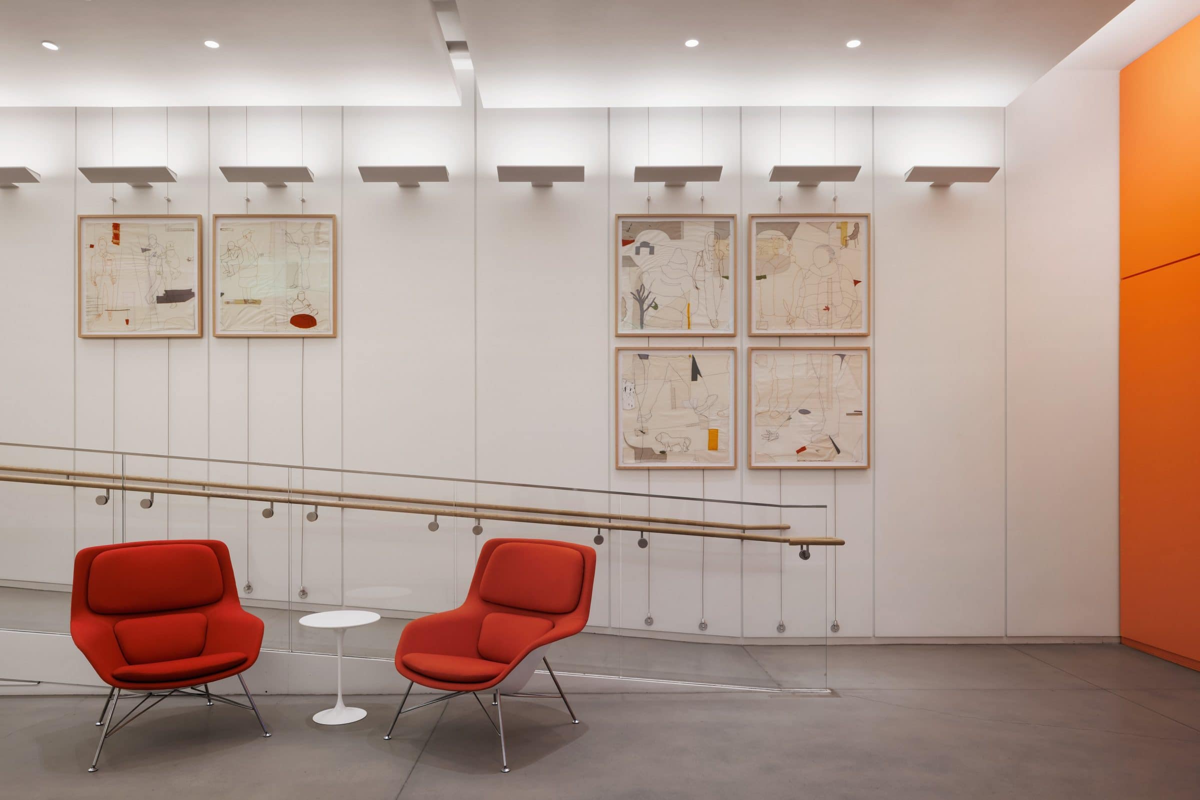Six framed stitched drawings on canvas hang in two groups on a white lobby wall behind two red lounge chairs
