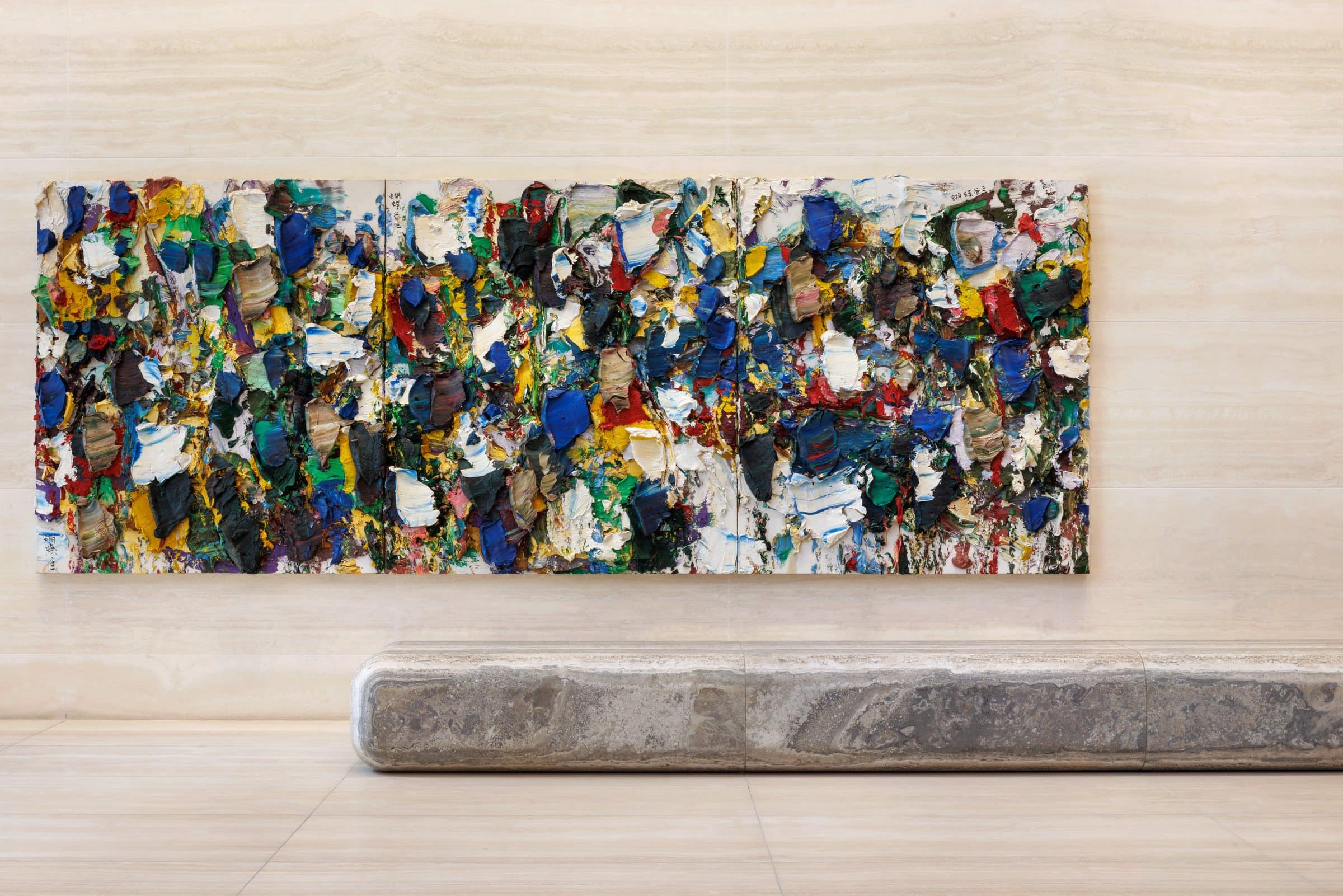 Frontal view of a long painting built from thick blue, yellow, green, red and white paint, above a stone bench