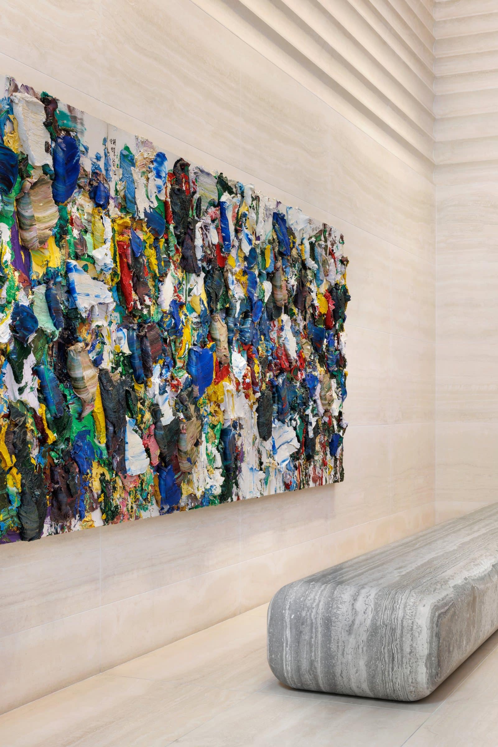 Angled close view of thick multicolored paint projecting from a long canvas above a gray stone bench