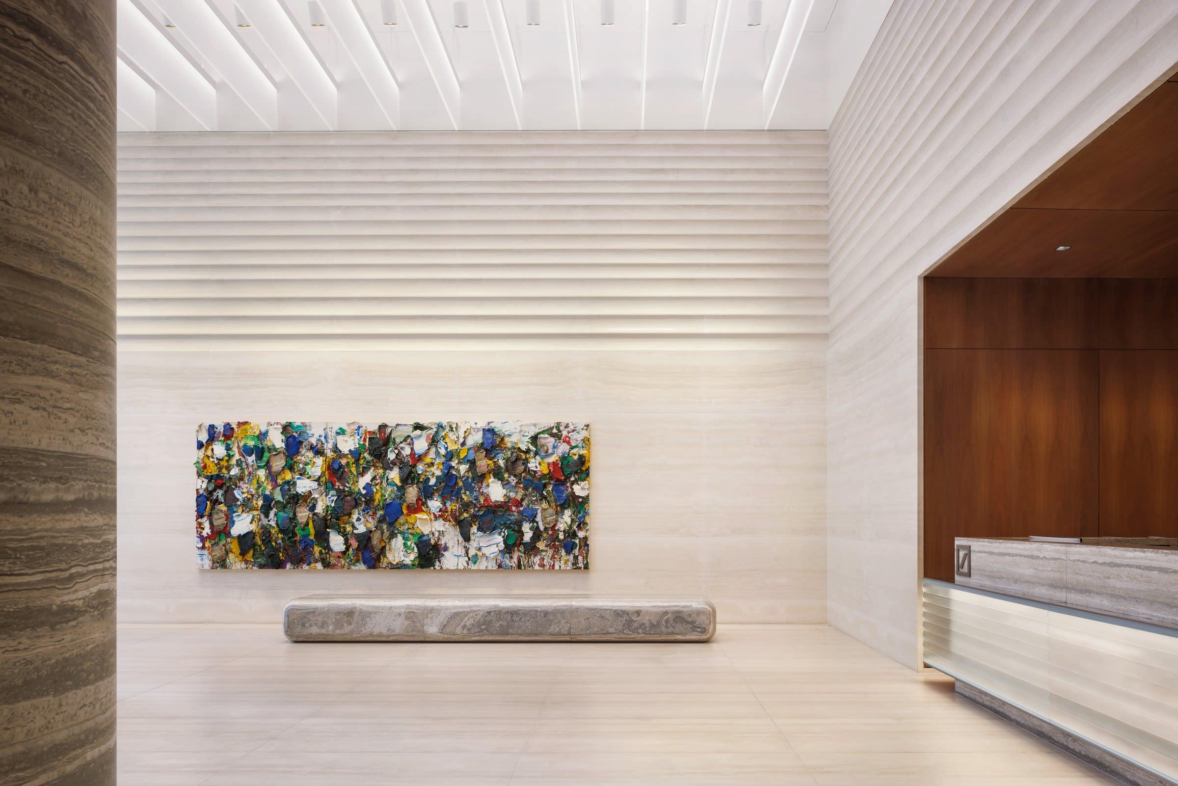 Wide frontal view across the lobby to a colorful impasto painting and stone bench, with reception at right