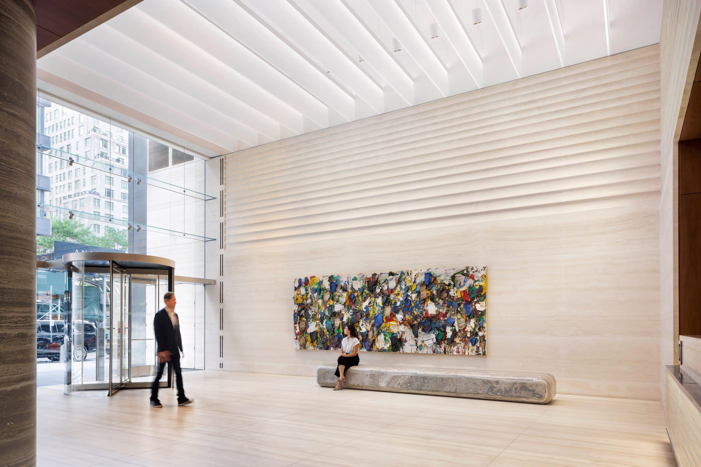 A visitor walks by the glass entrance while another sits below the long multicolored painting