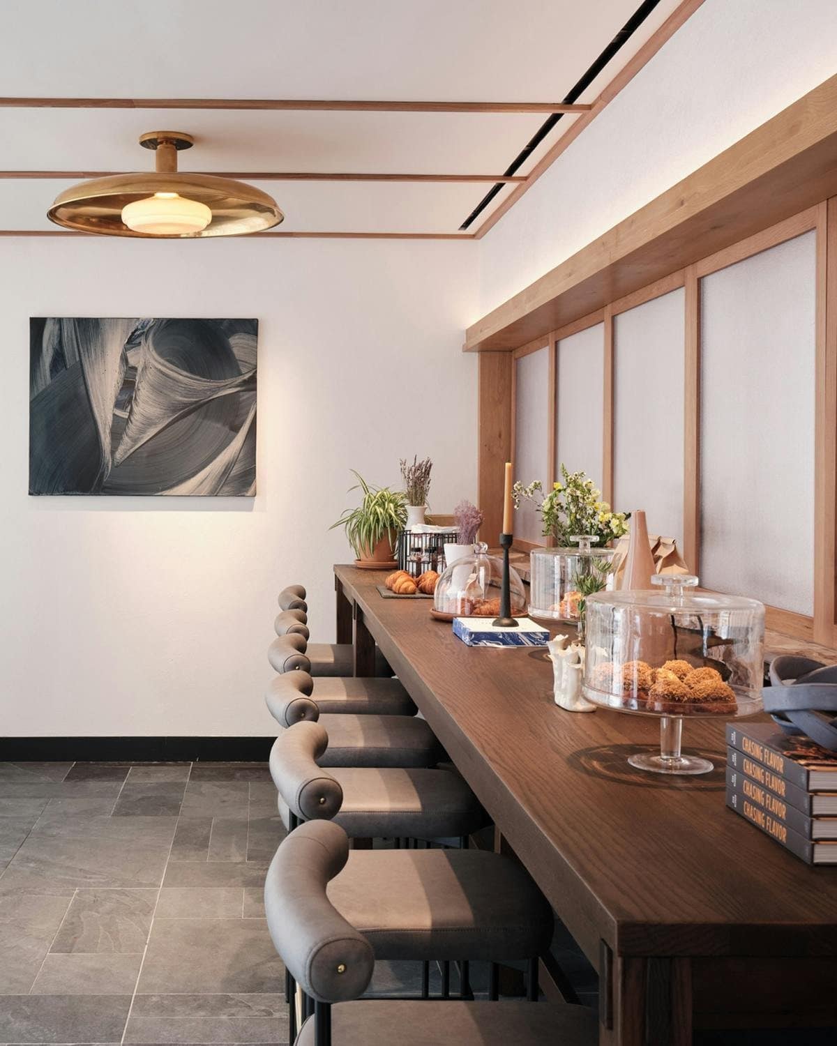 Bakery counter with pastries under glass domes, gray stools and a square blue-gray painting of sweeping brushstrokes