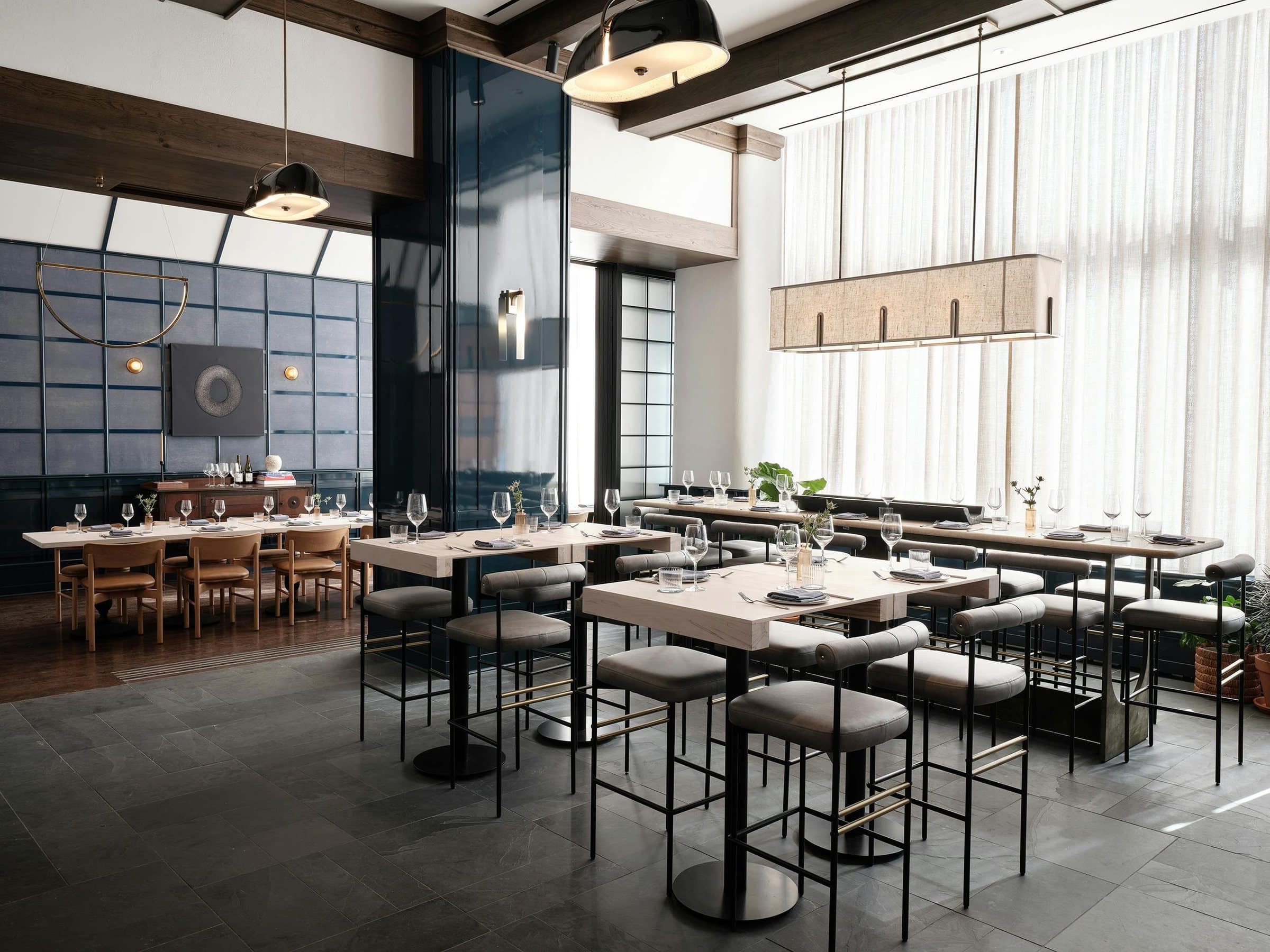 High-top dining area beneath a linen pendant, looking into a blue-paneled private dining room with a textured ring artwork