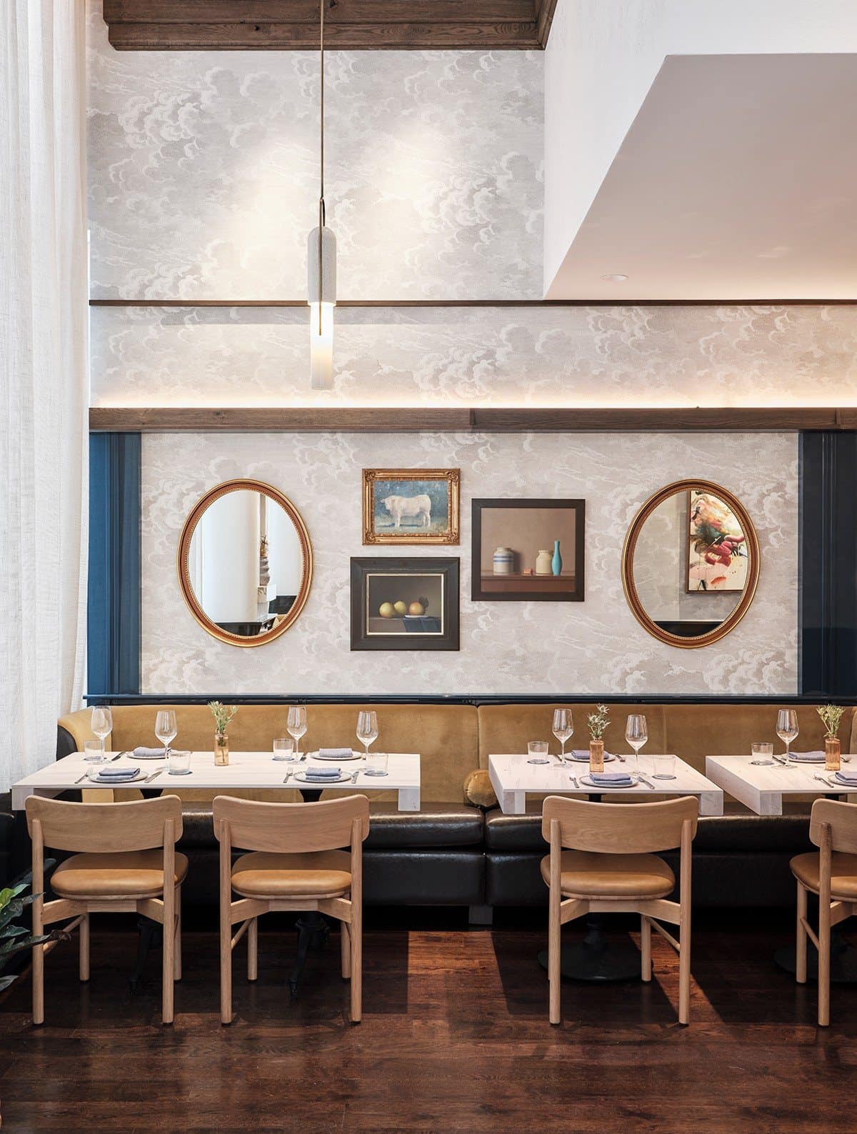 Greywind banquette beneath small paintings of a white cow, fruit and ceramic vessels, arranged between oval gold mirrors