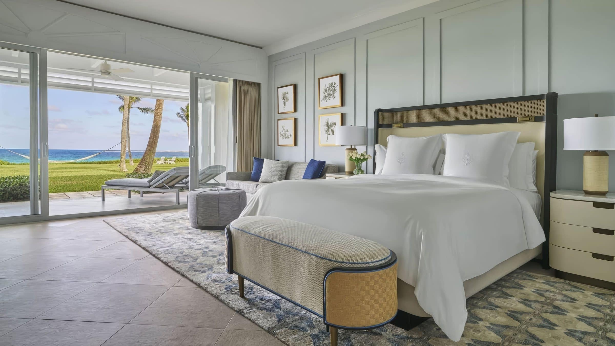 Ocean-view guest bedroom with a king bed and four small framed botanical works above a sofa beside the terrace doors