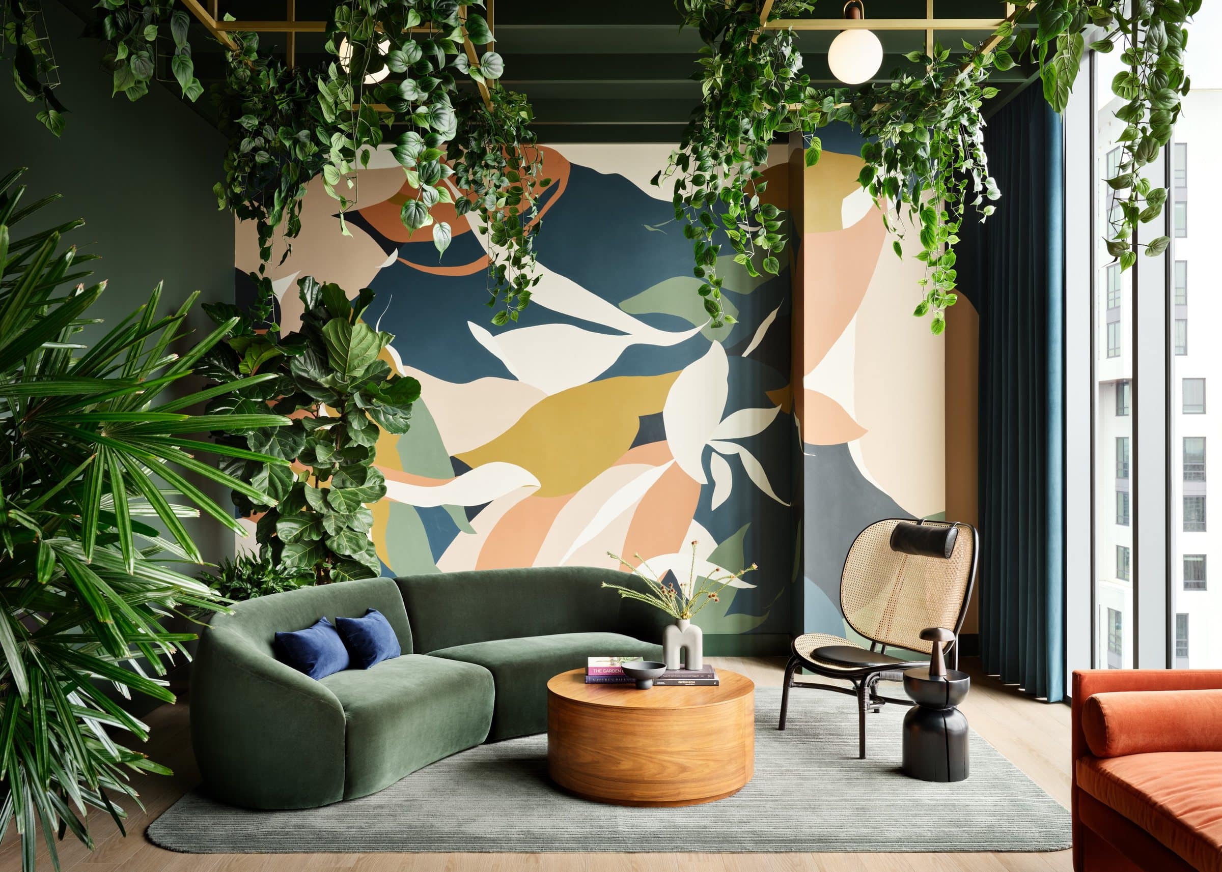 Plant-filled lounge with a curved green sofa in front of a large abstract mural in cream, yellow, peach and dark blue