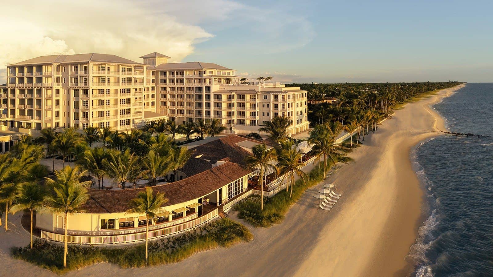 Aerial view of a cream resort building and a low clubhouse among palms beside a long sandy beach at sunset