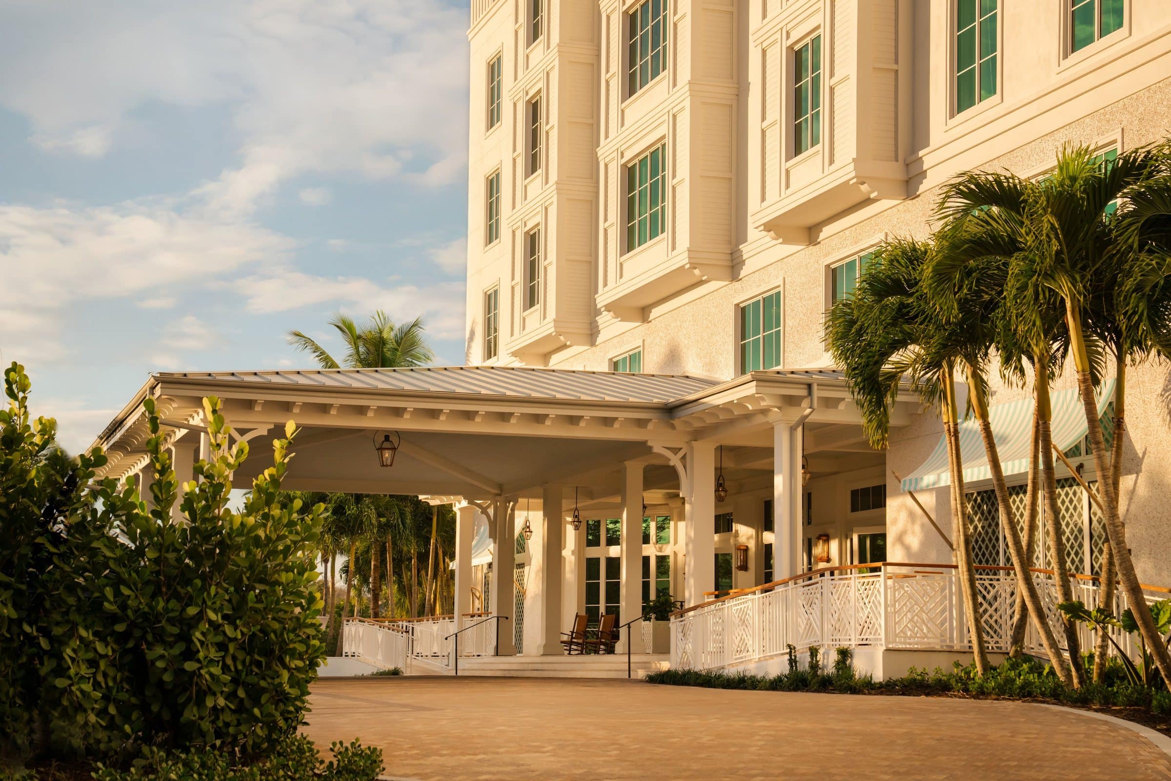 A white porte-cochère with lanterns and lattice railings below a cream building with green-framed windows, framed by palms