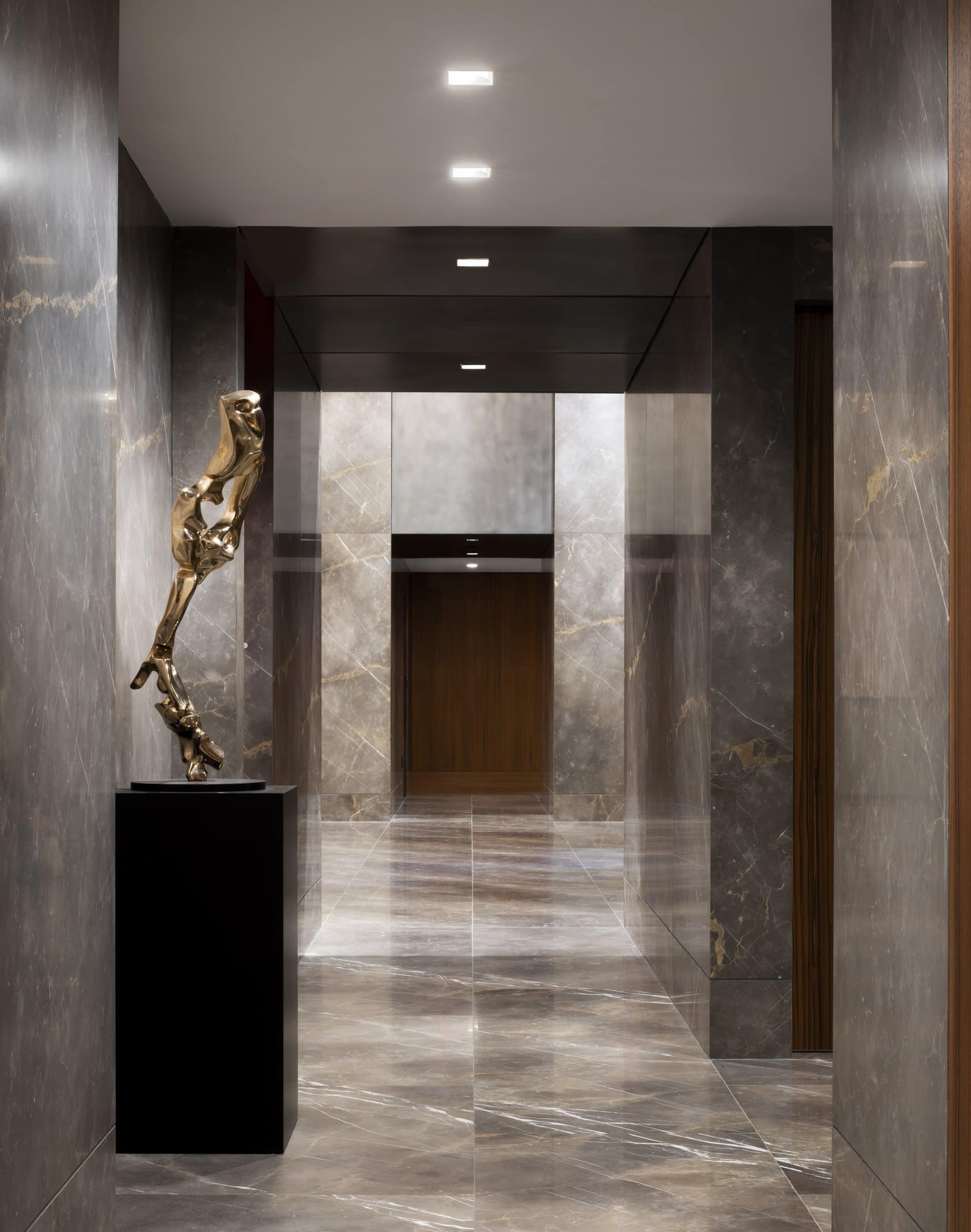 Tall irregular bronze sculpture on a black plinth beside a dark stone corridor