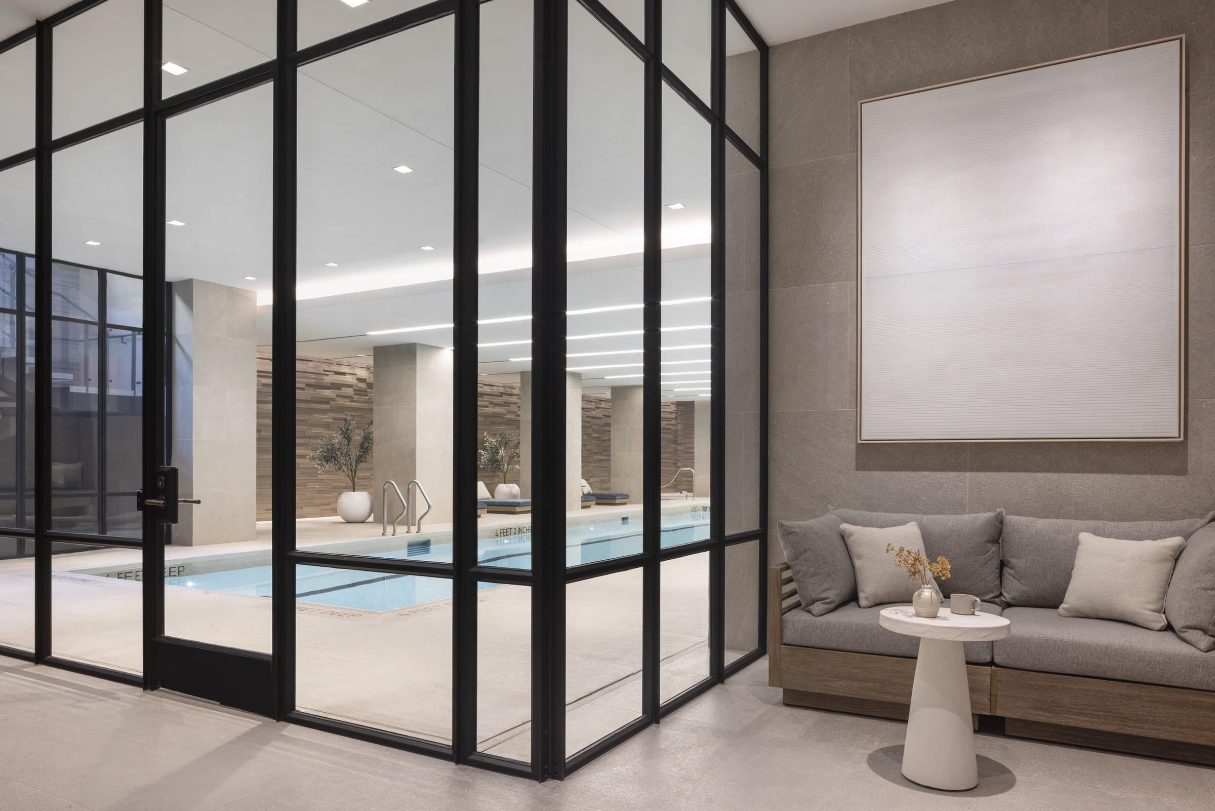 Indoor pool seen through glass doors beside a gray sofa and a large pale atmospheric work
