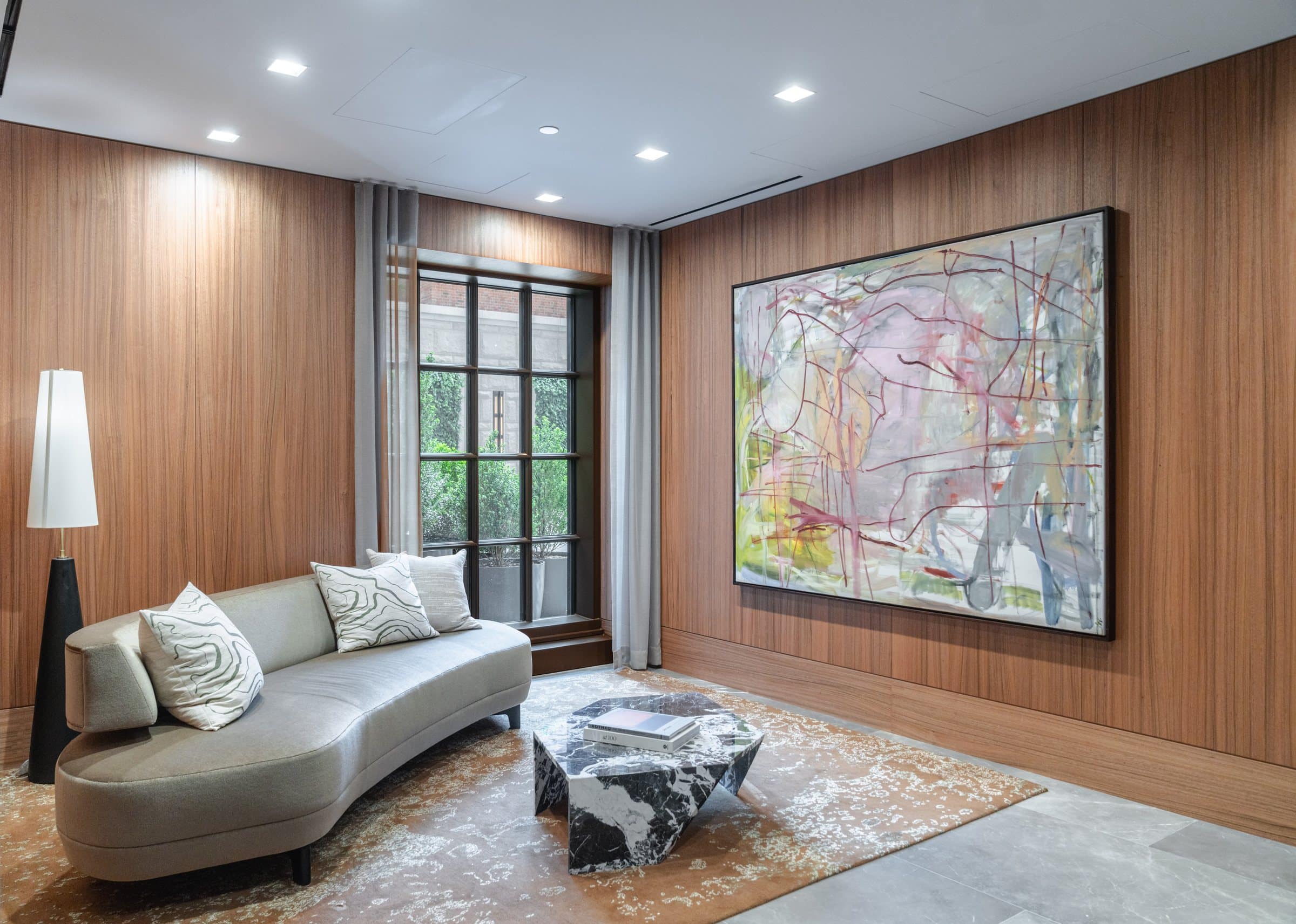 Pale pink, green and gray linear abstract painting above a curved sofa and marble table in a wood-paneled lounge