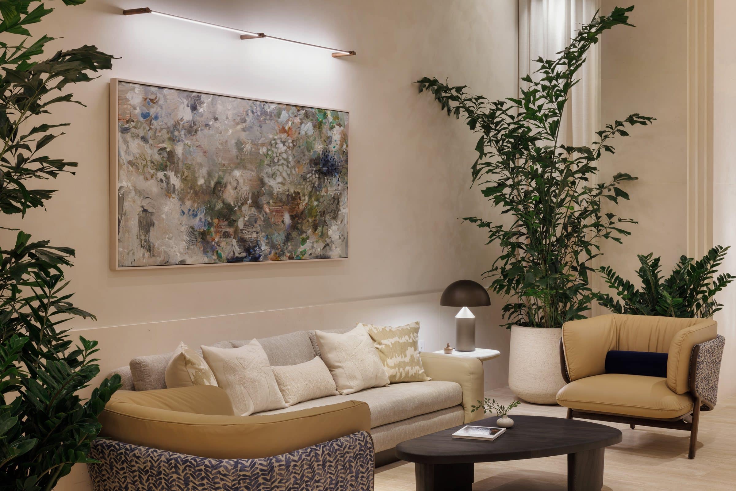A wide mottled gray, green and blue abstract painting above a cream sofa between tall plants