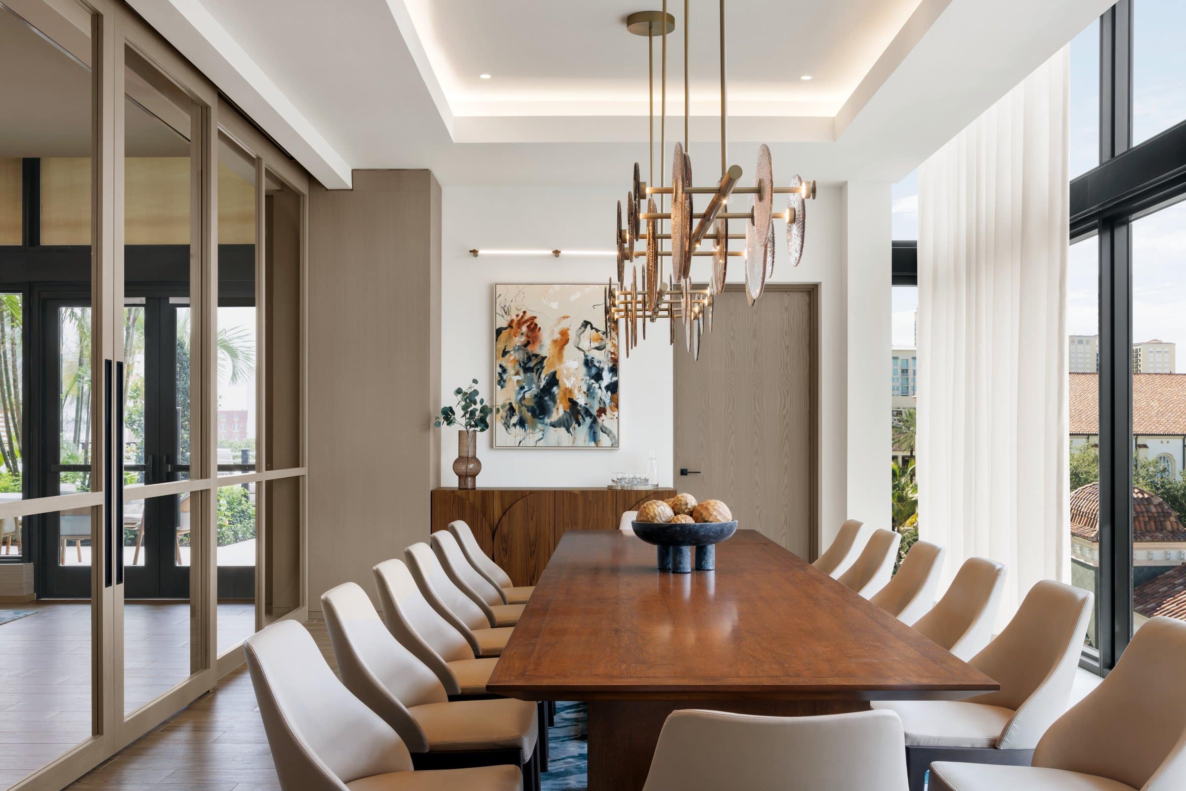 Long private dining table beneath a brass pendant, with a cream, orange and black gestural painting on the far wall