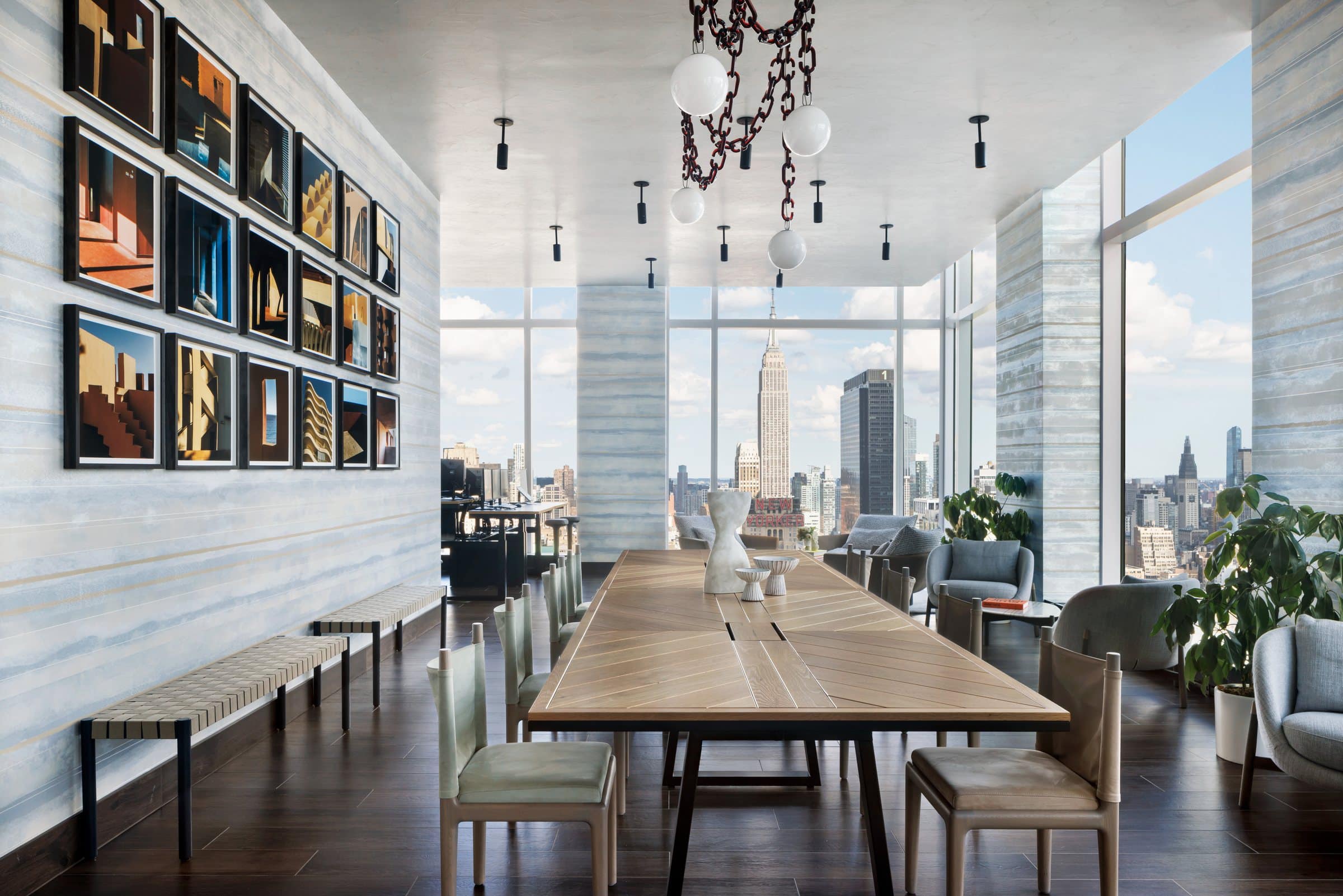 Long coworking table beneath a grid of architectural photographs, with floor-to-ceiling skyline views
